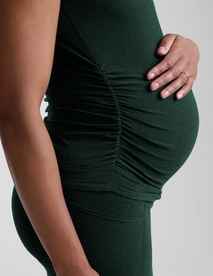 model woman wearing a dark green maternity t-shirt and leggings against a light gray background.