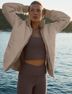 woman in a beige jacket and purple outfit standing by a body of water.