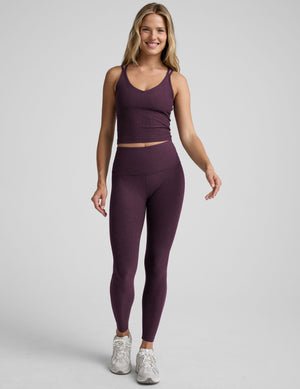 model is wearing a purple v-neck tank top with criss-cross strap back detail and purple high-waisted midi length leggings on a plain background.