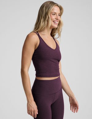 model is wearing a purple v-neck tank top with criss-cross strap back detail and purple high-waisted midi length leggings on a plain background.